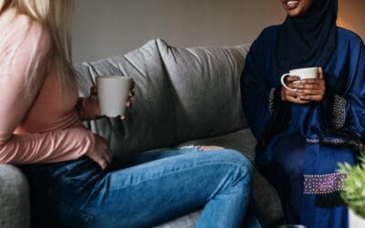 An american womann and a foreign woman are sitting together on a couch in a living room. They are enjoying conversation over a hot beverage.