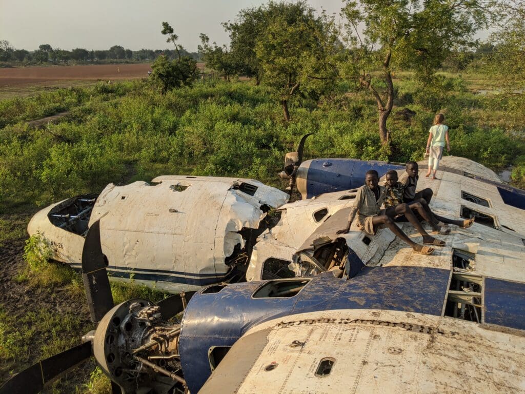 3 North African boys sit on the scraps of two prop plane while a little caucasian girl looks off into the distance of the green field and farmland.