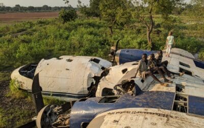 3 North African boys sit on the scraps of two prop plane while a little caucasian girl looks off into the distance of the green field and farmland.