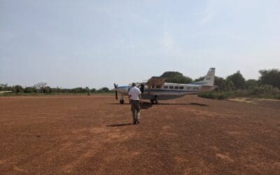 An East African pilot walks along the red dirt runway to a white, blue-striped, single prop plane.