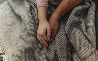 A husband and wife loosely holding hands over the covers.