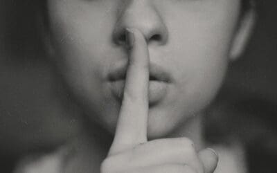 A close up portrait from the nose down of a young woman with her pointing finger over her lips. She's shushing someone.