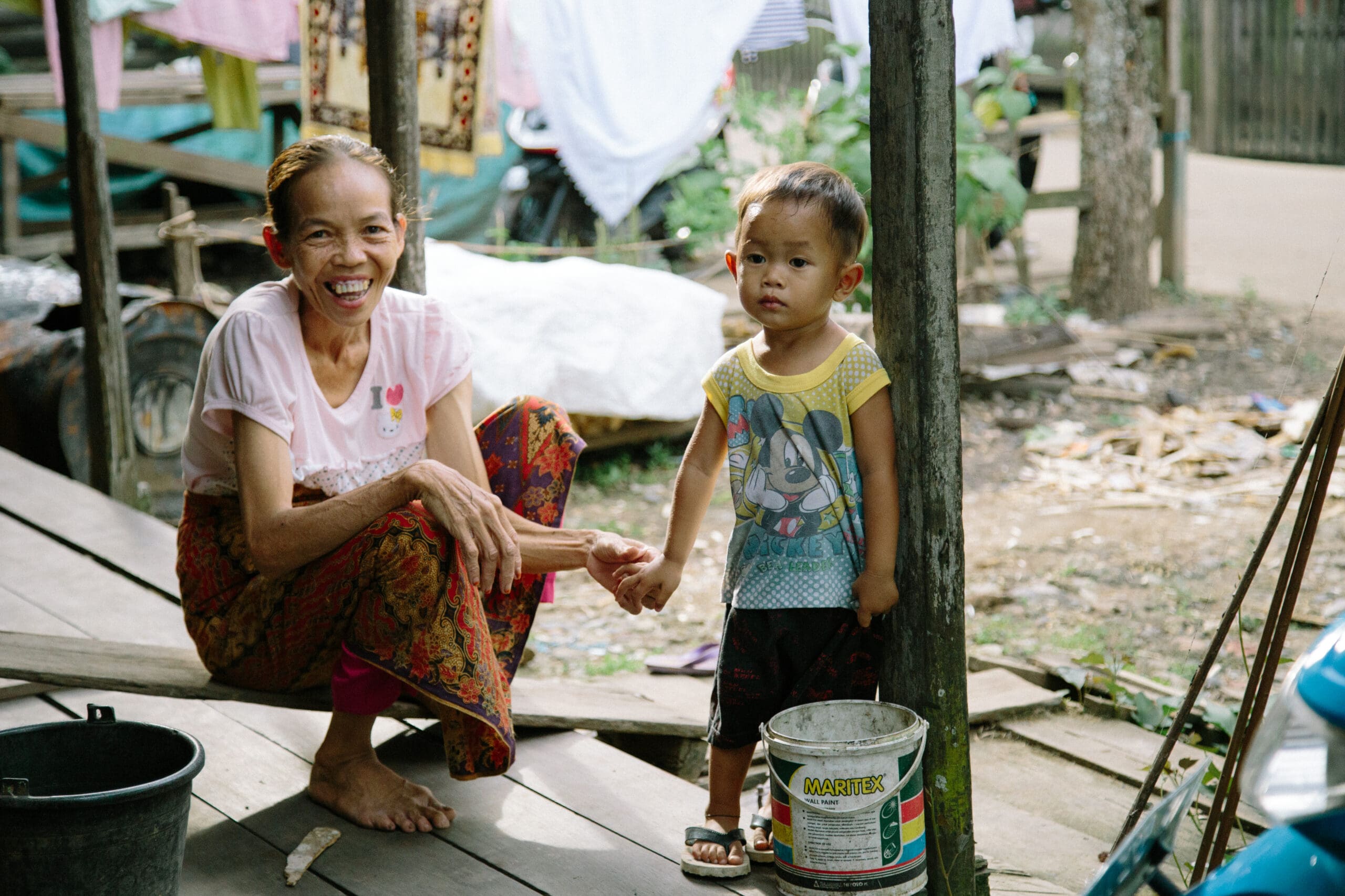 A smiling woman and a young child sit outside in a rural setting. The woman is wearing a light pink Hello Kitty shirt and colorful patterned pants, while the child wears a yellow shirt with a cartoon print. Greenery and a covered motorbike are visible in the background.